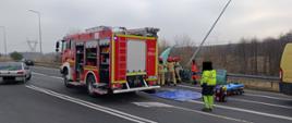Zdjęcie ukazuje szeroki kadr miejsca wypadku, gdzie strażacy PSP i ratownicy medyczni prowadzą działania ratunkowe. W tle widoczny jest pojazd strażacki z włączonymi światłami ostrzegawczymi oraz inne pojazdy biorące udział w zdarzeniu. Na pierwszym planie widać ratownika medycznego oraz sprzęt ratunkowy rozłożony na jezdni, w tym nosze i torbę medyczną. Strażacy zabezpieczają wrak pojazdu oraz prowadzą ewakuację poszkodowanych. Sytuacja wymaga dużego zaangażowania służb, co wskazuje na powagę zdarzenia.