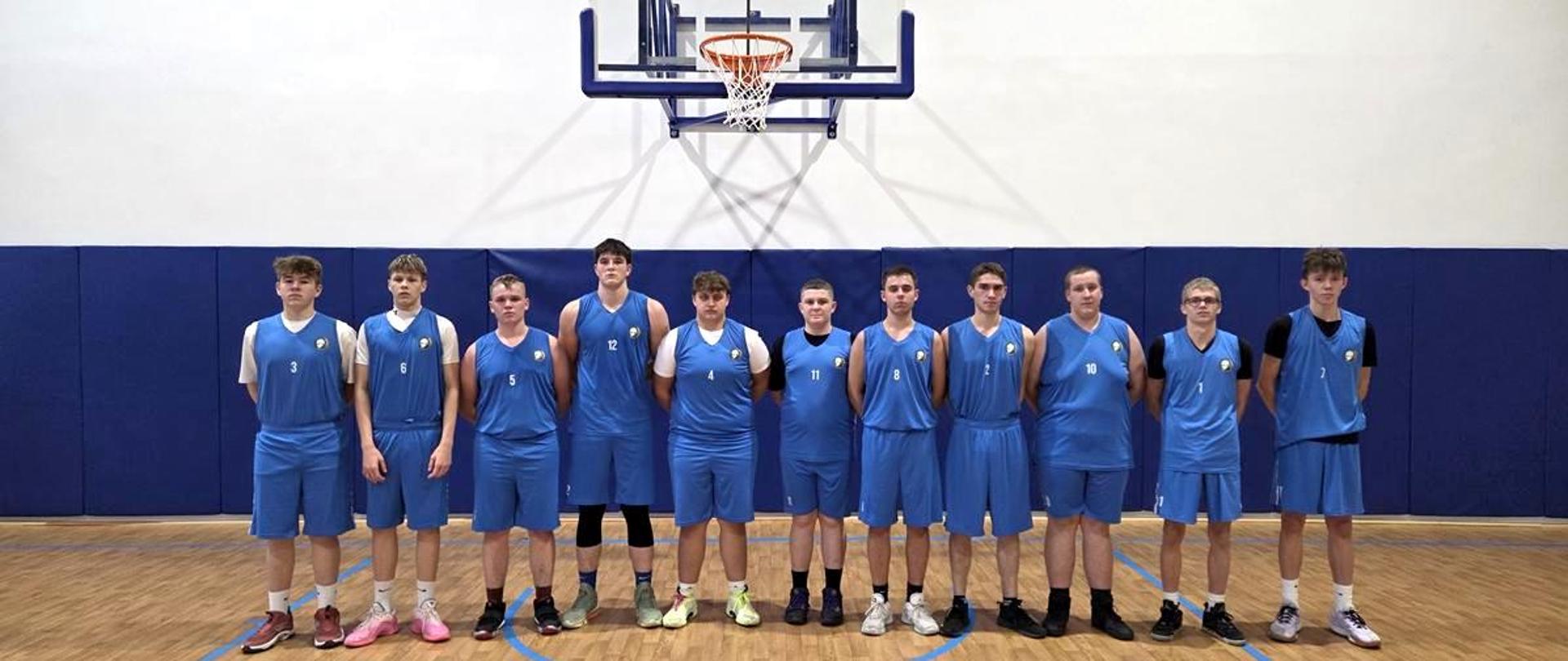 Drużyna koszykówki w niebieskich strojach, składająca się z jedenastu chłopców, stoi w szeregu na parkiecie hali sportowej przed koszem.
