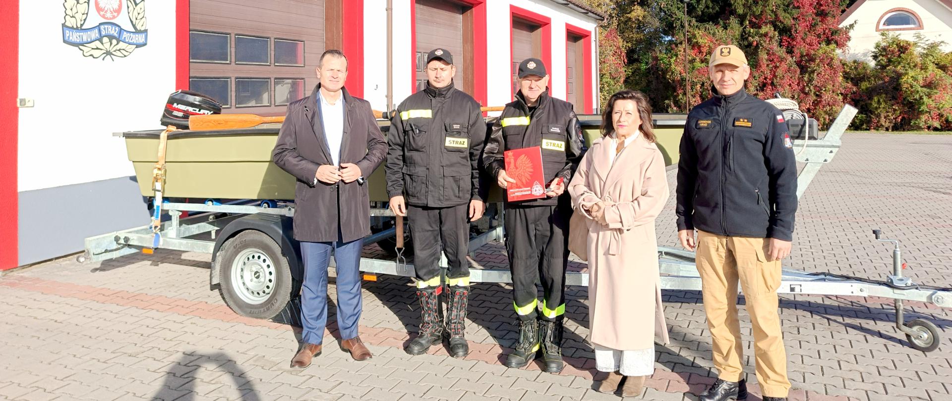 Na zdjęciu znajduje się pięć osób: od prawej strony stoi Zastępca Komendanta Powiatowego PSP w Nowym Dworze Mazowieckim brygadier Łukasz Kochanowski, z jego lewej strony stoi Pani Magdalena Biernacka Starostwa Nowodworski, kolejno z lewej Prezes OSP Cieksyn druh Andrzej Sosnowski i druh Damian Gruszka. Pierwsza osoba z lewej to Burmistrz Nasielska Pan Radosław Kasiak. Osoby te stoją przed łodzią na przyczepie, która została przekazana do OSP Cieksyn. W tle widać garaże Jednostki Ratowniczo-Gaśniczej w Nowym Dworze Mazowieckim z logo Państwowej Straży Pożarnej. Prezes OSP Cieksyn trzyma w ręku symboliczny dokument przekazania łodzi, który otrzymał w ramach przekazania. 