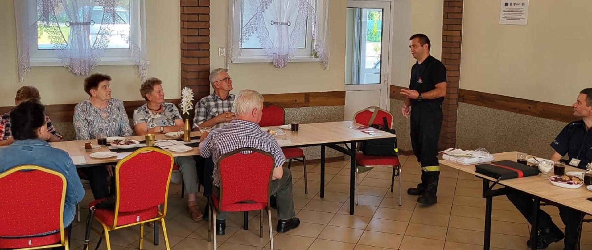 Na zdjęciu strażak omawiający zagadnienia związane z bezpieczeństwem seniorów. Przy stole siedzą uczestnicy spotkania - seniorzy. Zdjęcie zrobione na sali wiejskiej.