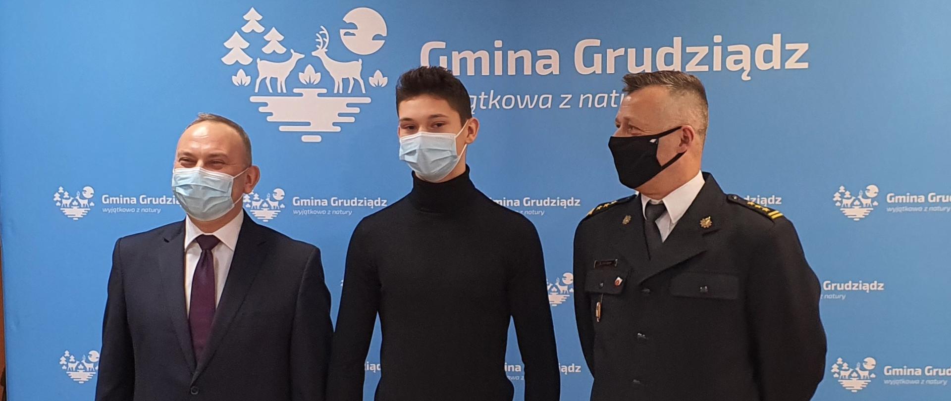 Zdjęcie przedstawia 3 mężczyzn stojących na tle niebieskiej ścianki z napisem Gmina Grudziądz wewnątrz pomieszczenia. Stojący po prawej ubrany jest w czarny mundur, środkowy w czarną bluzkę i spodnie, stojący po lewej w garnitur. 