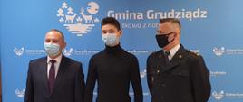 Zdjęcie przedstawia 3 mężczyzn stojących na tle niebieskiej ścianki z napisem Gmina Grudziądz wewnątrz pomieszczenia. Stojący po prawej ubrany jest w czarny mundur, środkowy w czarną bluzkę, stojący po lewej w garnitur. 