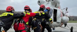 Na zdjęciu widzimy grupę ratowników w specjalistycznym umundurowaniu, którzy przygotowują się do wejścia na pokład śmigłowca. Mają na sobie kombinezony ochronne w kolorach czarnym i czerwonym, kaski ochronne z napisem "PSP" (Państwowa Straż Pożarna), uprzęże oraz sprzęt alpinistyczny .
Na zdjęciu widać, że śmigłowiec stoi na płycie lotniska lub innego placu manewrowego, a ratownicy trzymają się za ramiona, co jest typową procedurą bezpieczeństwa przy wchodzeniu do maszyny z działającym wirnikiem.