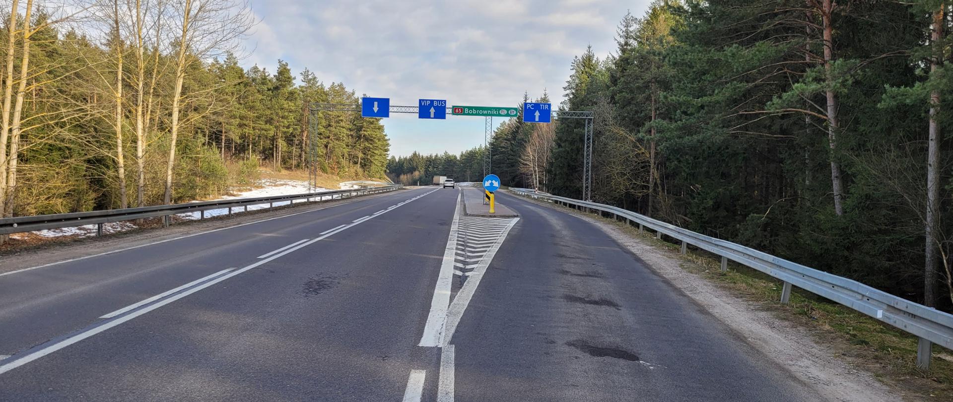 Droga prowadząca do przejścia granicznego w Bobrownikach.
W oddali widoczne pojazdy, znaki kierunkowe nad drogą.
