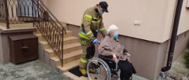 Strażak prowadzi wózek inwalidzki, na którym siedzi starsza kobieta w berecie. W tle budynek i schody do budynku.
