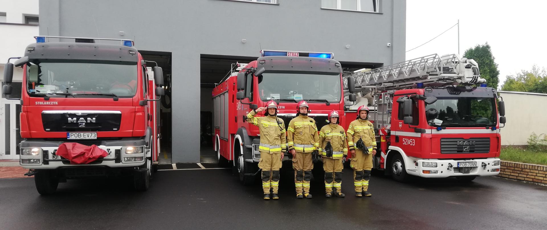 strażacy oddają honor na tle pojazdów strażackich