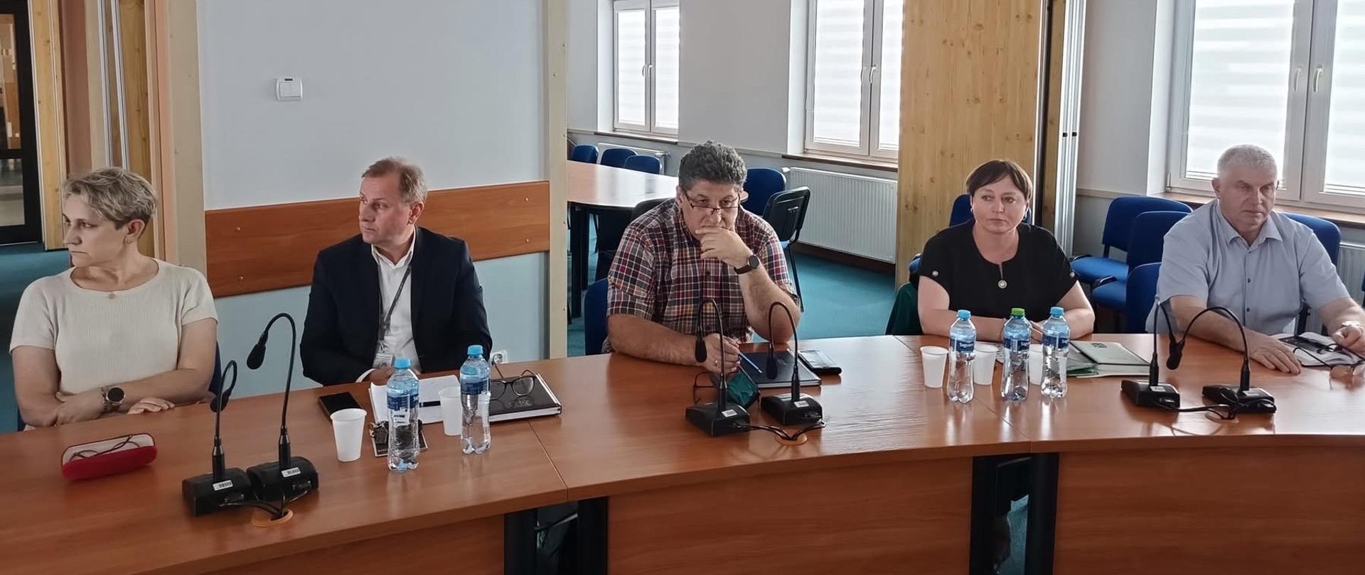 Grupa ludzi siedząca przy stole. Trzech mężczyzn i dwie kobiety. Na stole są mikrofony oraz woda w butelkach. Pomieszczenie jest jasne. 