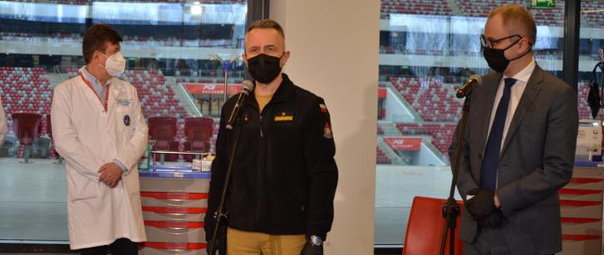 strażak stoi przed mikrofonem, obok stoi mężczyzna w białym fartuchu oraz drugi mężczyzna w garniturze