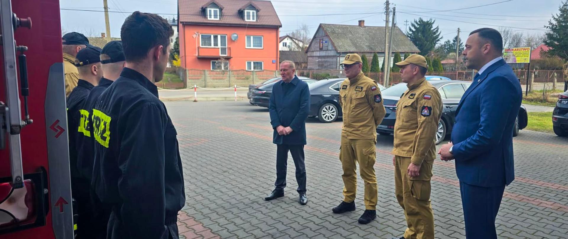 Grupa strażaków w czarnych mundurach koszarowych stoi w szeregu naprzeciwko delegacji urzędników i Zastępcy Komendanta Głównego PSP. Wiceminister oraz dowódcy PSP prowadzą rozmowę. Spotkanie odbywa się na placu przed remizą, w tle widoczne są budynki mieszkalne oraz zaparkowane samochody służbowe.
