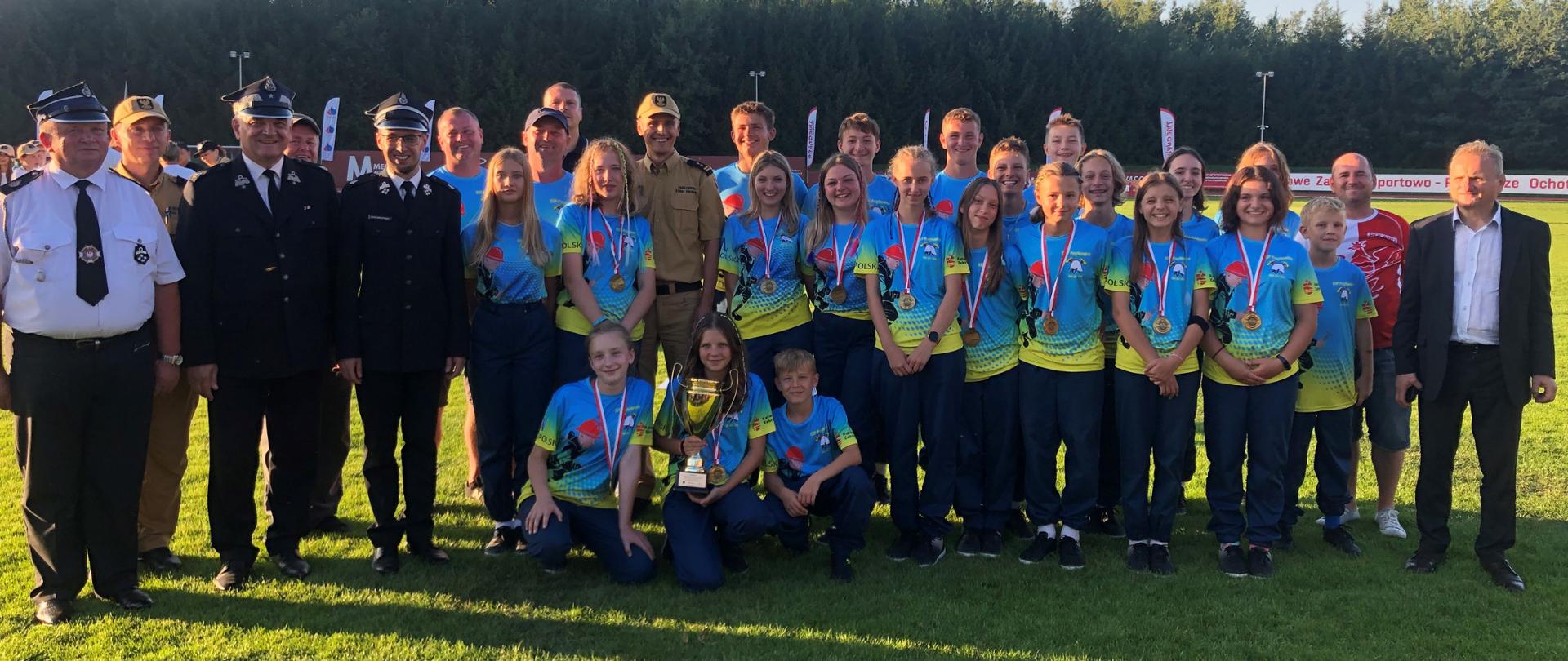 Widok z przodu. Zdjęcie grupowe drużyny MDP OSP Przytkowice wraz z opiekunami, Prezesem Zarządu Głównego Związku OSP RP, Zastępcą Małopolskiego Komendanta Wojewódzkiego PSP, Zastępcą Komendanta Powiatowego PSP w Wadowicach, Prezesem Oddziału Powiatowego ZOSP RP w Wadowicach.