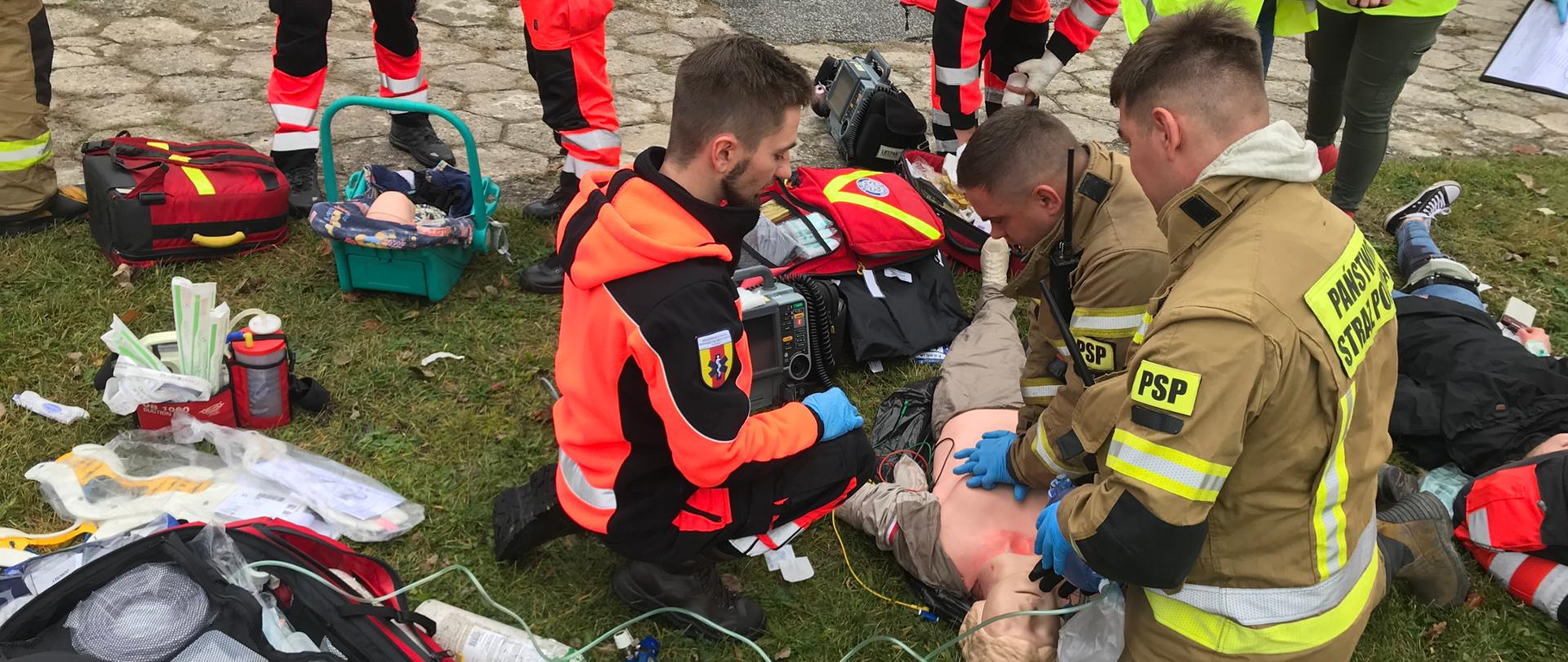 Dwóch strażaków i ratownik medyczny symulujący udzielanie kwalifikowanej pomocy przedmedycznej na fantomie szkoleniowym