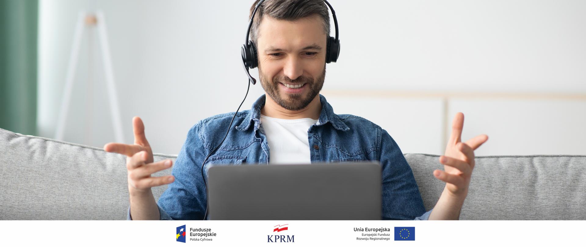 Zdjęcie mężczyzny siedzącego na kanapie i korzystającego z laptopa. Na uszach ma słuchawki z mikrofonem, bierze udział w wideospotkaniu. Uśmiecha się, w geście zadowolenia unosi ręce w stronę ekranu komputera.