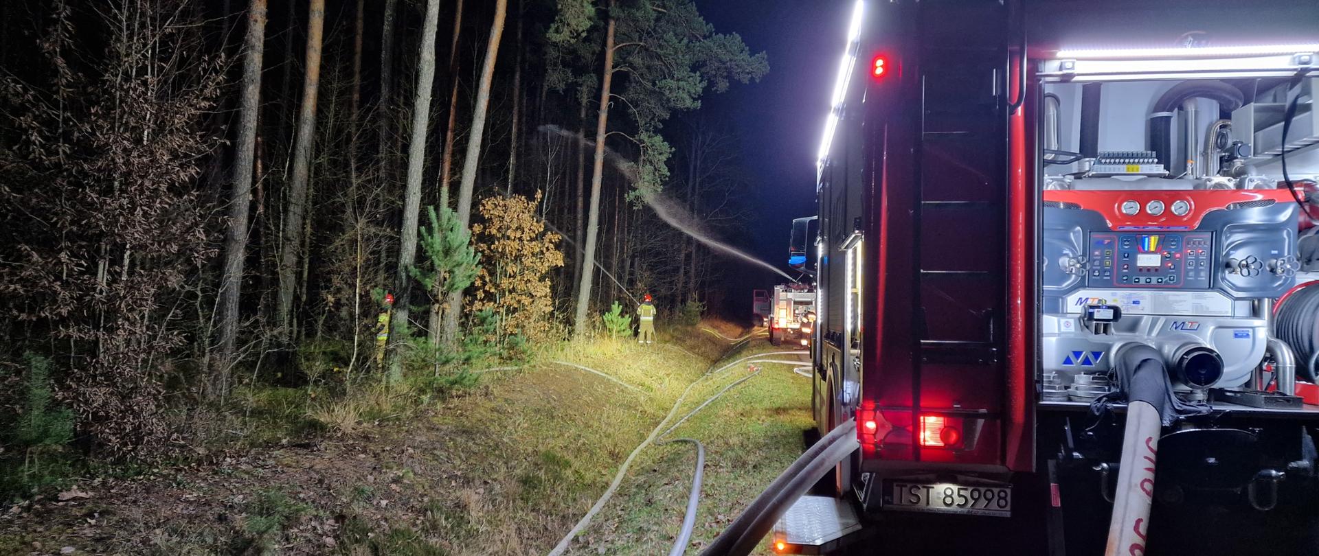 Samochód pożarniczy z podpiętymi liniami gaśniczymi, w tle strażacy podający prądy gaśnicze. Pora nocna