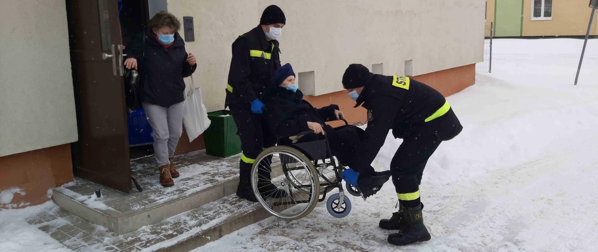 Dwóch strażaków znoszących kobietę na wózku inwalidzkim z bloku mieszkalnego.