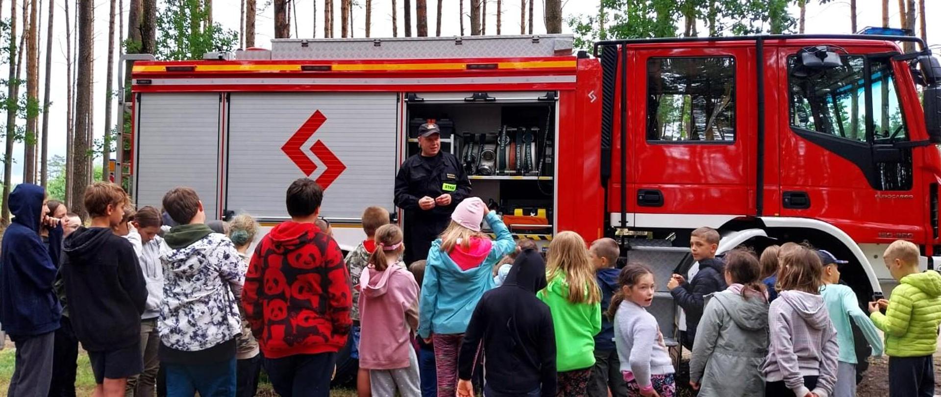 Strażak stoi przed grupą dzieci. Strażak ma na sobie umundurowanie koloru czarnego. Za strażakiem stoi wóz strażacki. Wóz strażacki jest koloru czerwonego z białymi, szarymi, czarnymi i żółtymi elementami. Dzieci mają na sobie kolorowe ubrania. Wszyscy znajdują się w lesie. W tle widać drzewa. 