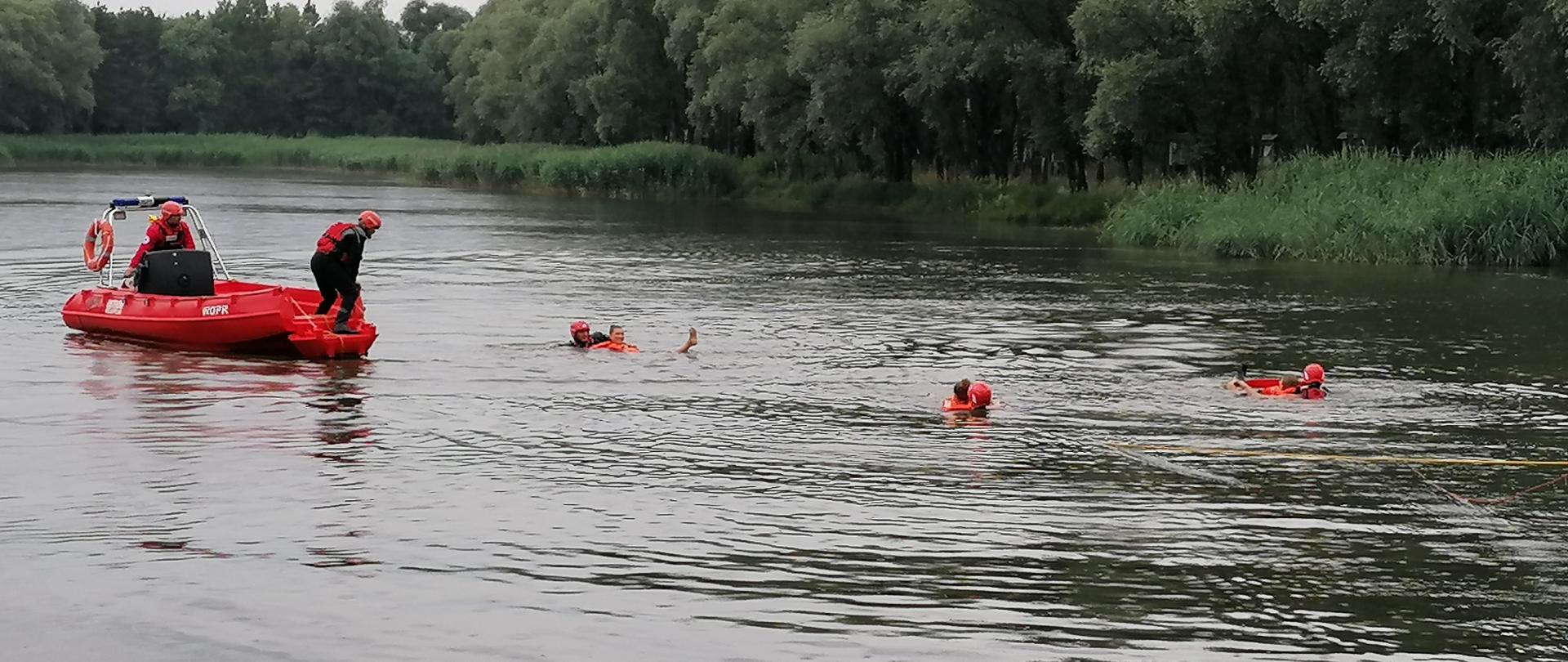 Zbiornik wodny, ratownicy PSP i WOPR podejmują 3 osoby poszkodowane w wodzie. Dwóch ratowników WOPR znajduje się na łódce. W tle las.