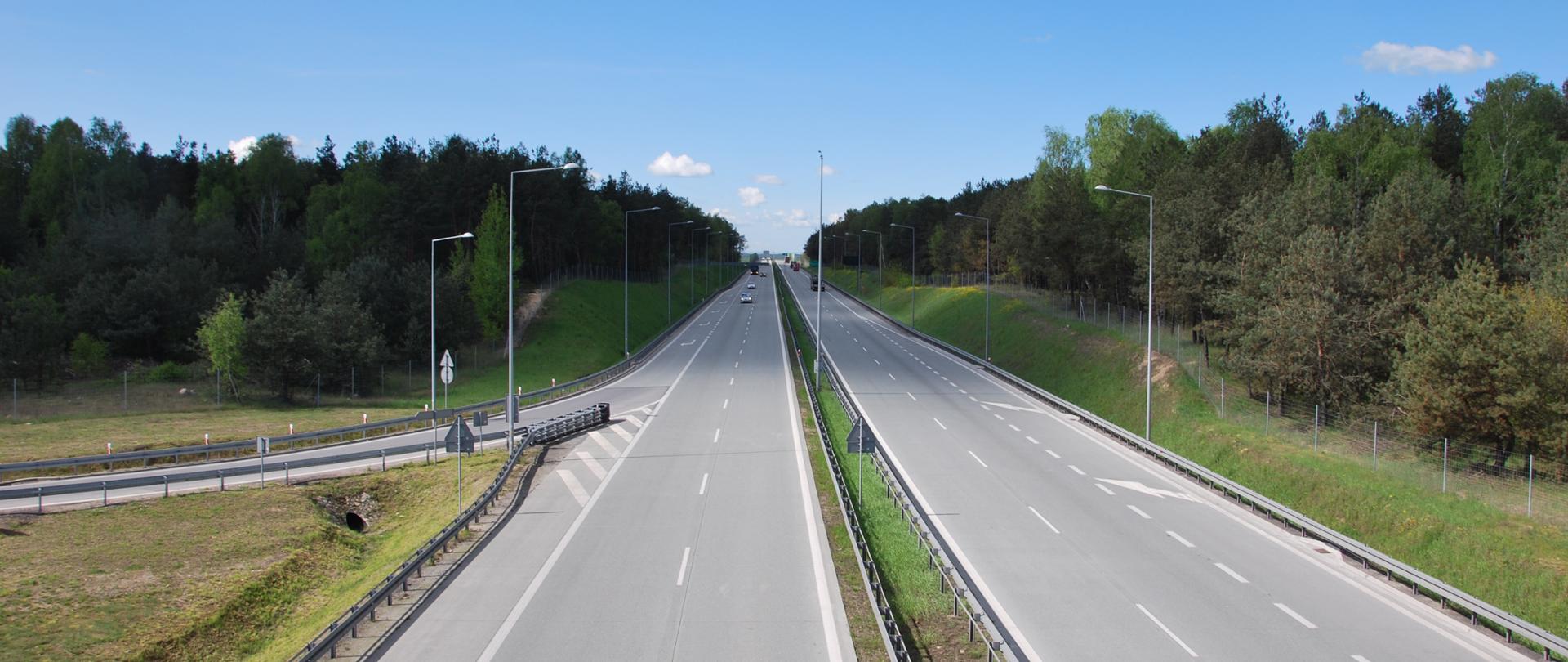 Droga ekspresowa S8. Dwujezdniowa droga ekspresowa o dwóch pasach ruchu przedzielona pasem zieleni. Po obu stronach drogi rośnie las. Z prawej strony zdjęcia widać zjazd z drogi ekspresowej.