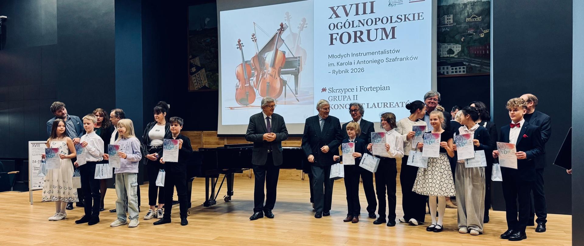 Na zdjęciu widać grupę młodych uczestników stojących na scenie podczas uroczystości wręczenia dyplomów. Za nimi znajduje się ekran z napisem „XVIII Ogólnopolskie Forum Młodych Instrumentalistów im. Karola i Antoniego Szafranków, Rybnik 2026”. Obok dzieci stoją dorośli, jurorzy konkursu i organizatorzy. W tle widoczny jest fortepian, a uczestnicy trzymają w rękach dyplomy, uśmiechając się do zdjęcia.
