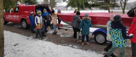Grupa dzieci znajduje się przy samochodzie Państwowej Straży Pożarnej Nissan Navara do którego podczepiona jest łódka. Strażacy pokazują dzieciom sprzęt, który wykorzystują podczas działań ratownictwa wodno-lodowego. Wszyscy znajdują się nad stawem w Miejskim Ośrodku Sportu i Rekreacji w Rypinie.