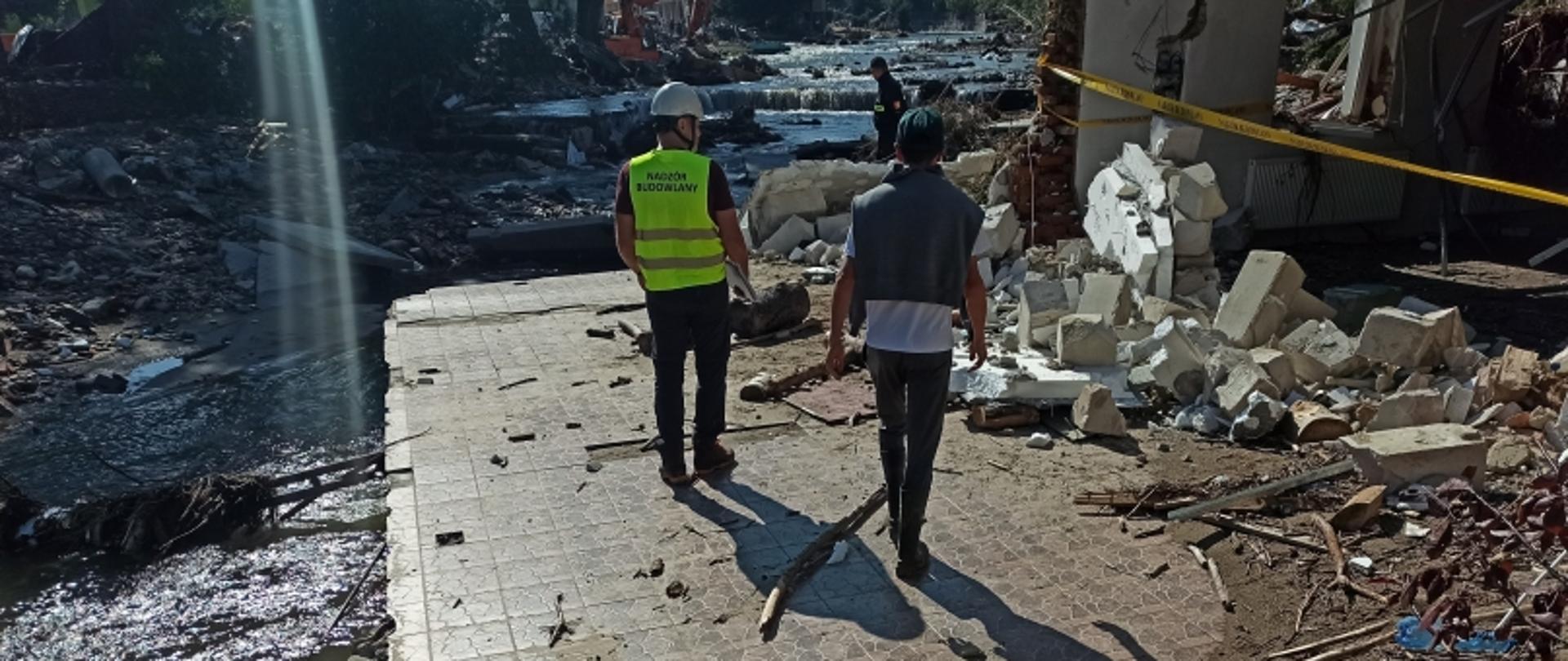 Dwóch ludzi na terenie zniszczeń po katastrofie – inspektor z napisem „NADZÓR BUDOWLANY”, gruz, zniszczenia, rzeka i taśma ostrzegawcza.