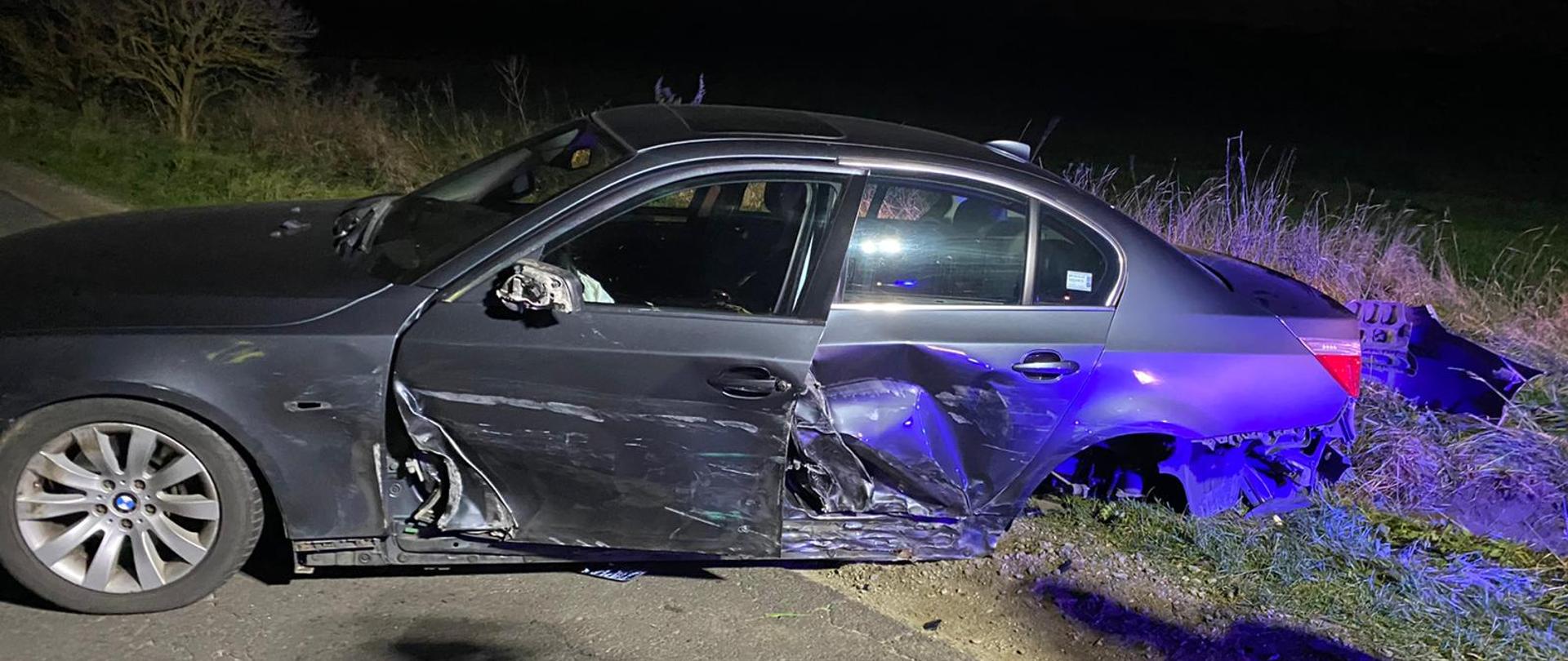 Widać czarny uszkodzony samochód od strony kierowcy, drzwi lekko otwarte, jest ciemno, brak tylnego koła