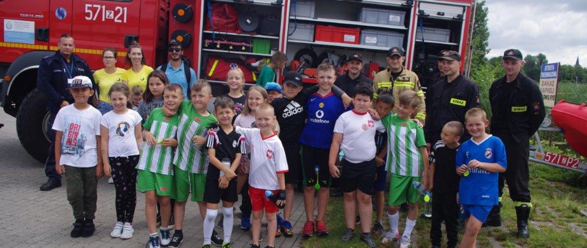 Zdjęcie przedstawia grupę dzieci, strażaków, policjanta na tle samochodu pożarniczego 