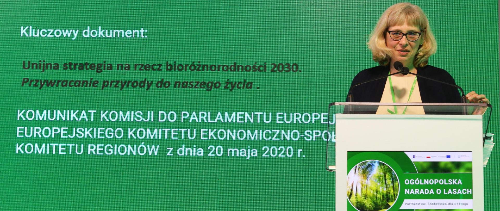 Na tle ściany ze slajdem prezentacji za mównicą stoi kobieta. Kobieta mówi do mikrofonu.