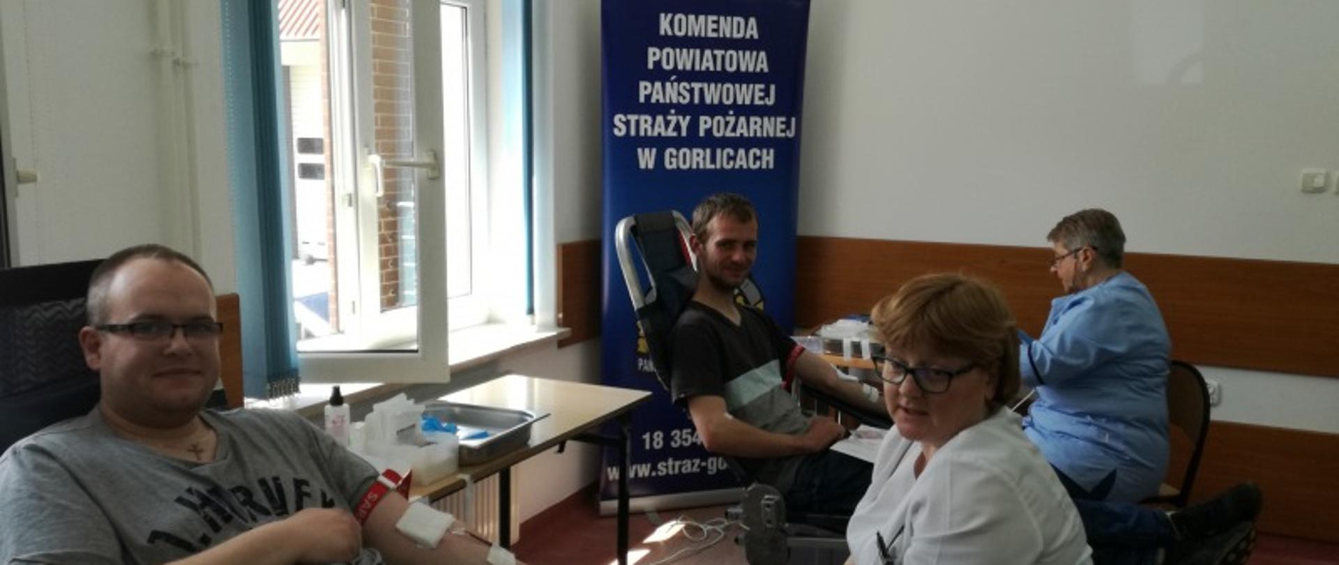 Na zdjęciu widoczne dwie osoby siedzące na fotelach w pomieszczeniu oddające krew przy nich pielęgniarki z Regionalnego Centrum Krwiodawstwa i Krwiolecznictwa z Oddziału Terenowego w Gorlicach