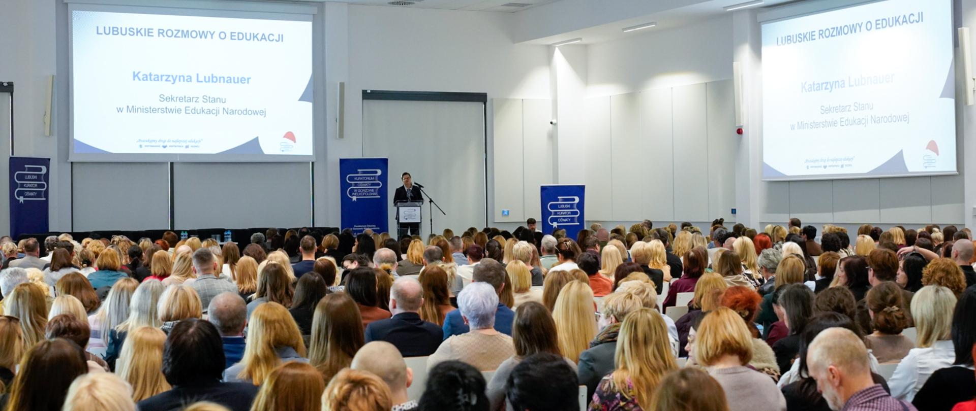 Szerokie ujęcie z tyłu sali konferencyjnej, wypełnionej dużą publicznością. Na scenie stoi prelegent, a za nim wyświetlane są dwa duże ekrany z napisem: "LUBUSKIE ROZMOWY O EDUKACJI" oraz "Katarzyna Lubnauer, Sekretarz Stanu w Ministerstwie Edukacji Narodowej".