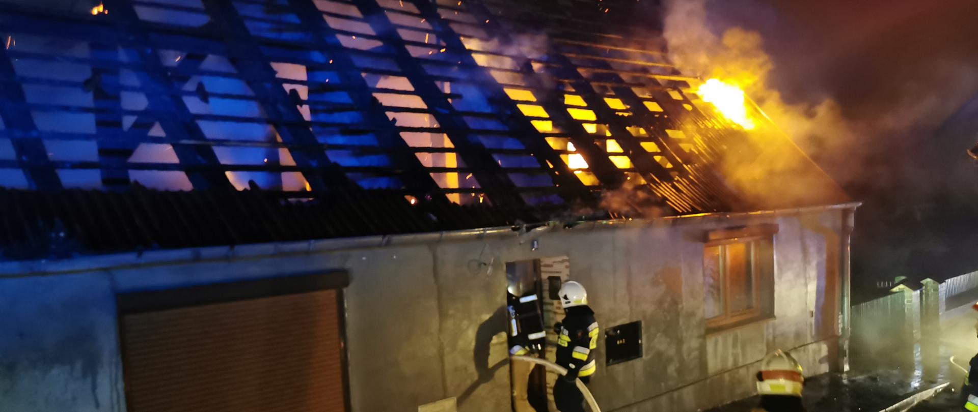 Zdjęcie przedstawia pożar dachu w budynku jednorodzinnym, widoczne płomienie na dachu budynku, strażacy wchodzą do środka obiektu z linią gaśniczą.