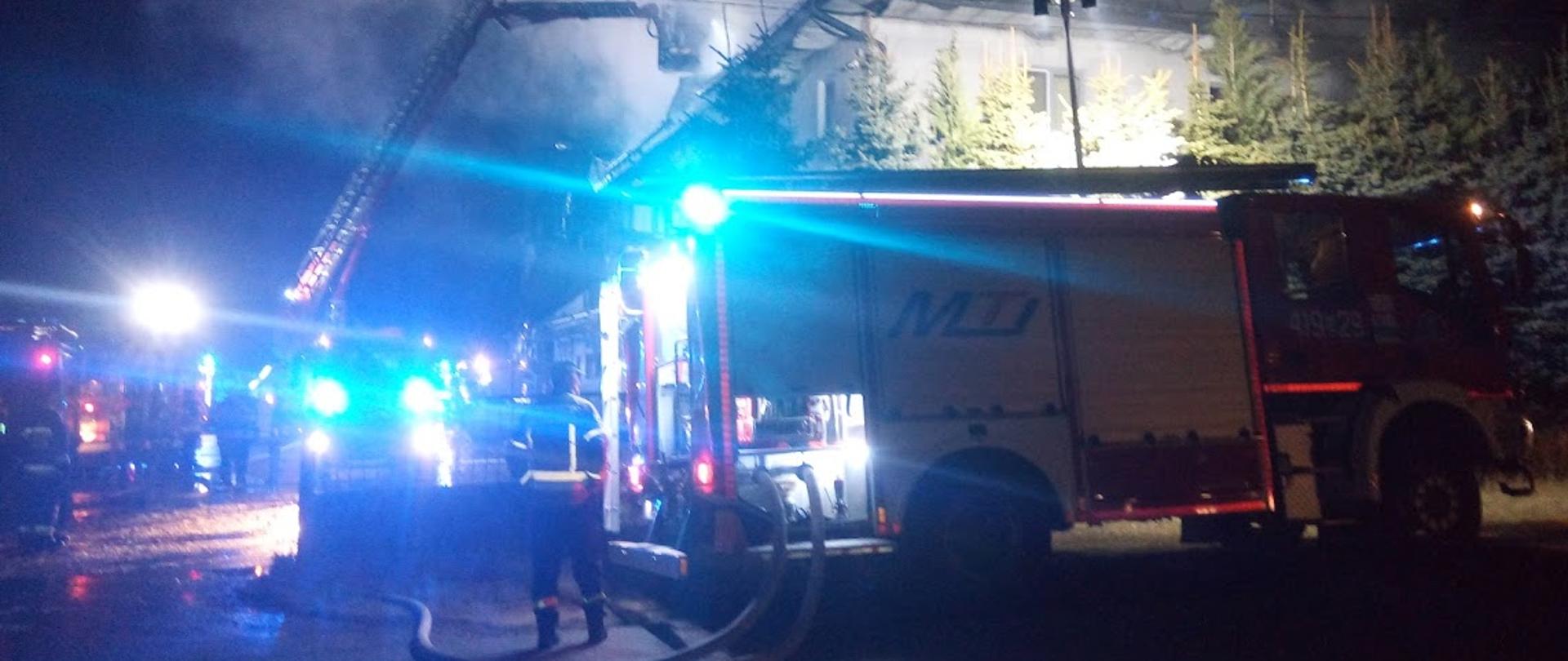 Zdjęcie przedstawia wozy ratowniczo-gaśnicze podczas akcji gaśniczej z włączonymi niebieskimi światłami 