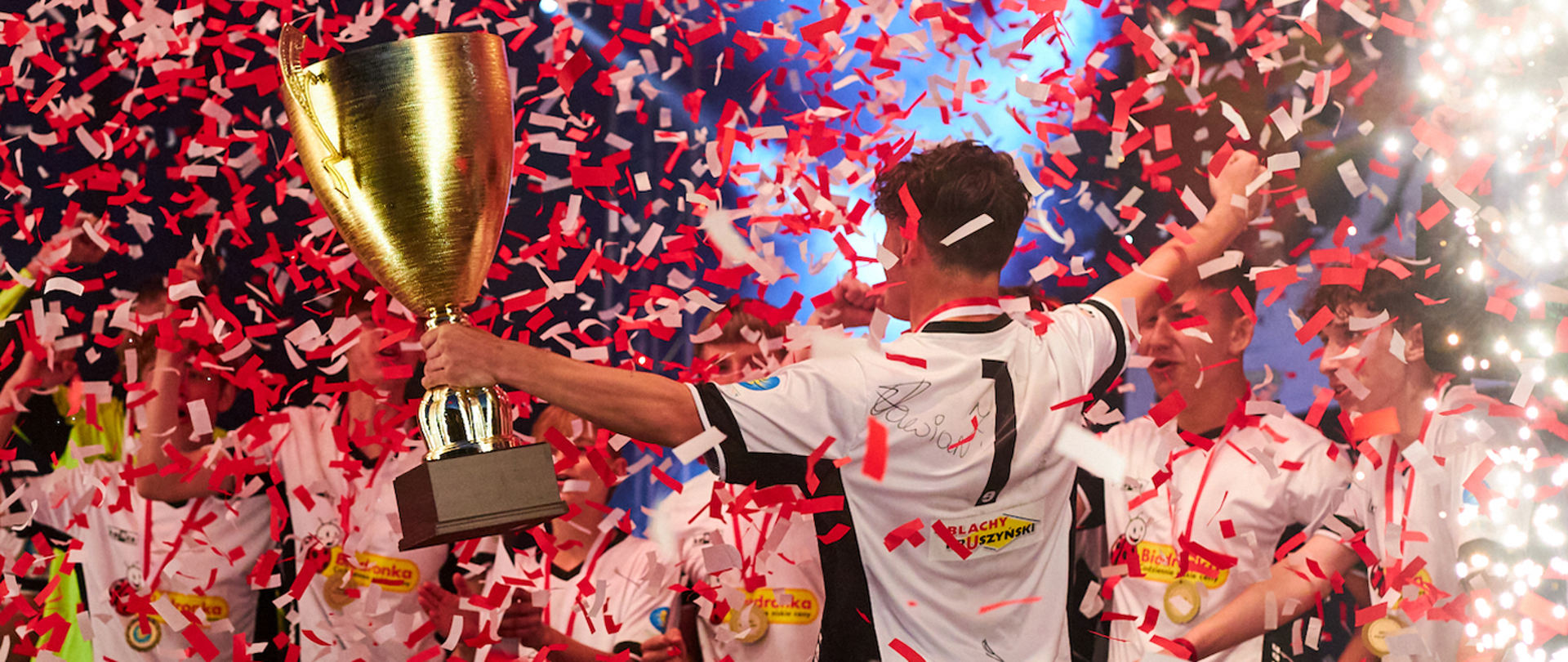 Dzieci świętują wygraną. Chłopic w białej koszulce z numerem 1 na plecach stoi tyłem do aparatu. W lewej ręce trzyma puchar. Z nieba leci biało-czerwone konfetti. W tle widać inne dzieci.