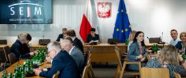 W sali przy podłużnych stołach siedzą ludzie, pod ścianą za stołem siedzi kobieta w czarnej marynarce, za nią pod ścianą na której wisi godło flagi Polski i UE, z boku na ścianie wisi monitor z napisem Sejm RP.