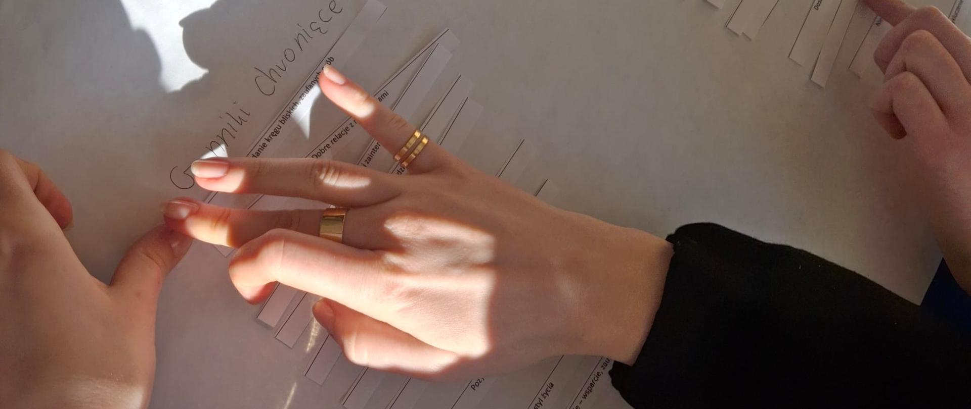 Zbliżenie na dłonie dwóch osób pracujących nad dużym arkuszem papieru. Na arkuszu znajdują się nagłówki „Zdrowie Psychiczne”, „Czynniki chroniące” oraz „Czynniki ryzyka”. Uczestnicy przyklejają paski papieru z nazwami poszczególnych zjawisk.