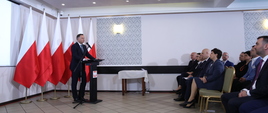 Na zdjęciu po lewej stronie prezydent Andrzej Duda na tle biało-czerwonych flag przy mównicy, po prawej siedzą uczestnicy konferencji.