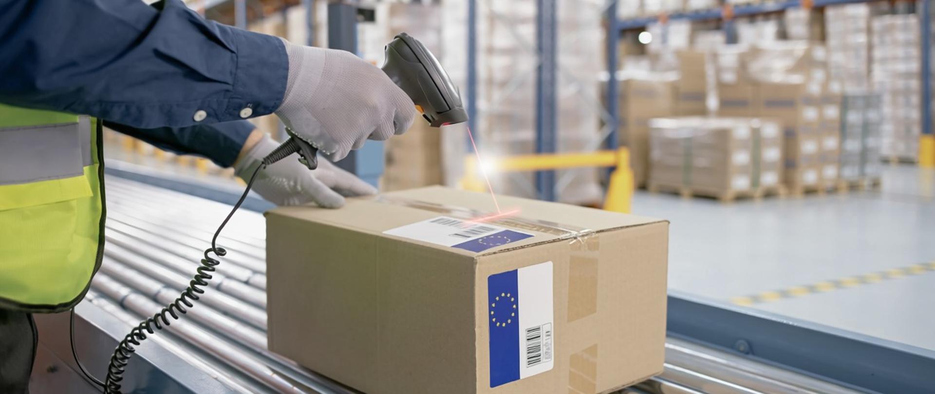 Pracownik magazynu skanuje kod kreskowy na kartonowym pudełku znajdującym się na taśmie transportowej w nowoczesnym centrum logistycznym. W tle widoczne są regały magazynowe z paletami.