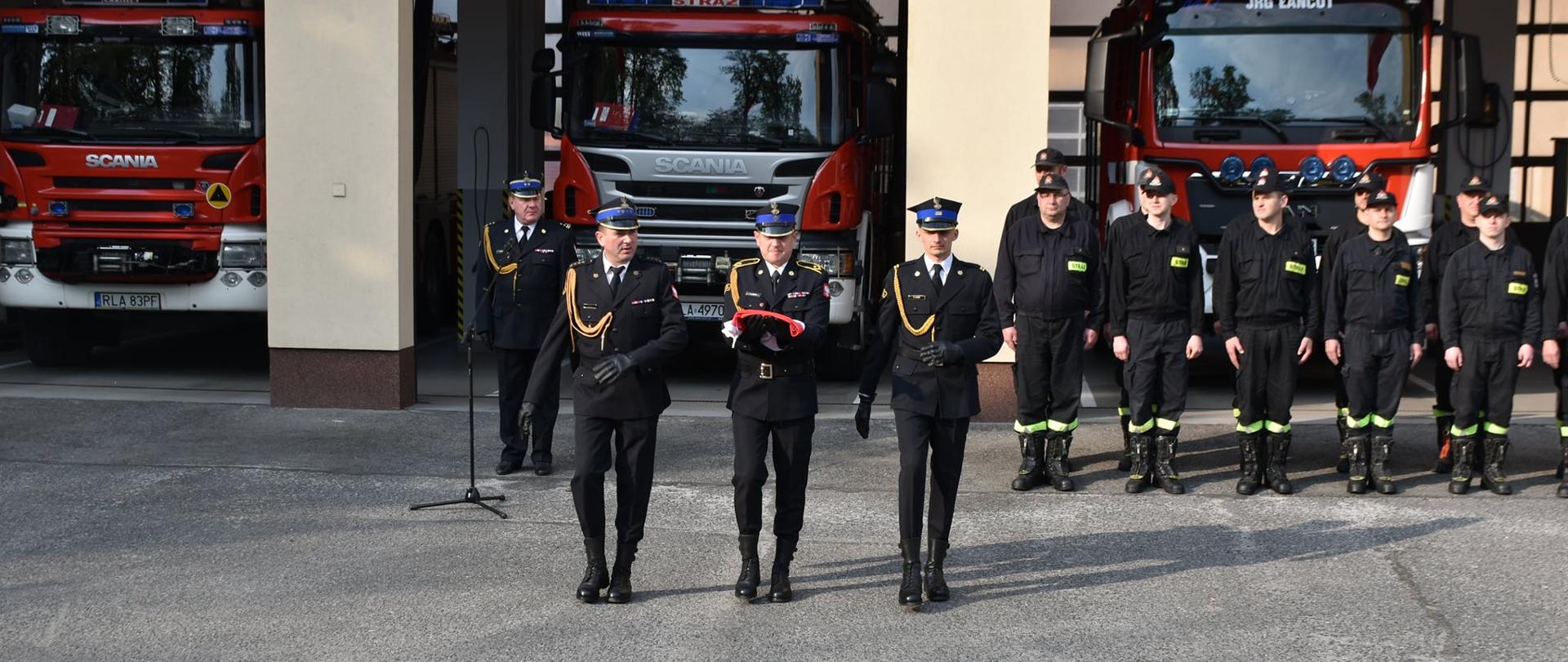 Strażacy w umundurowaniu wyjściowym ustawieni na uroczystej zbiórce przed garażem. Jeden ze strażaków trzyma złożoną biało -czerwoną flagę. Za srażakami ustawione samochody pożarnicze.