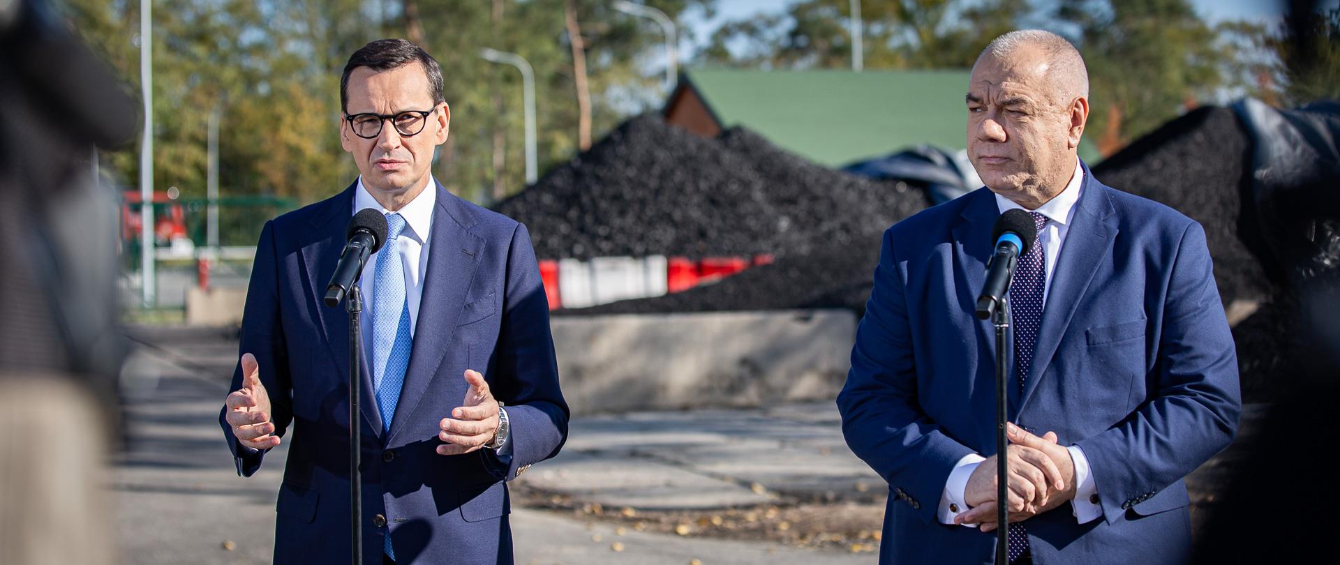Na zdjęciu premier Mateusz Morawiecki i wicepremier Jacek Sasin stoją za mikrofonami. W tle węgiel. 