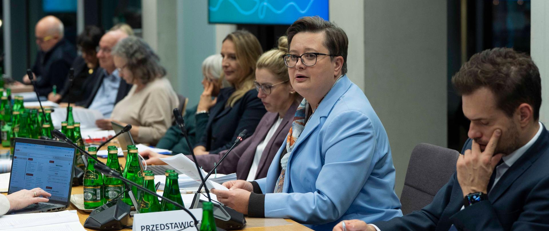 Zbliżenie na kobietę (Katarzyna Lubnauer) w jasnoniebieskiej marynarce i okularach, siedzącą przy stole z dokumentami i mikrofonem, prawdopodobnie wygłaszającą przemówienie lub prezentację podczas posiedzenia. W tle widoczny jest ekran z napisem "KOMPAS JUTRA".