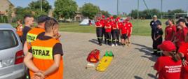 Obóz survivalowy w Sobiałkowie. Zdjęcie przedstawia 2 grupy dziewcząt i chłopców stojących na placu. Większość z nich ma na sobie czerwone czapki oraz koszulki. Obok srebrnego samochodu stoi 3 mężczyzn. Dwóch z nich ma założone kamizelki odblaskowe z napisem "STRAŻ".Trzeci to strażak PSP - w czarnej koszulce. Na środku placu znajduje się żółta deska ortopedyczna i czerwona torba medyczna.. W tle boisko i inni uczestnicy obozu.