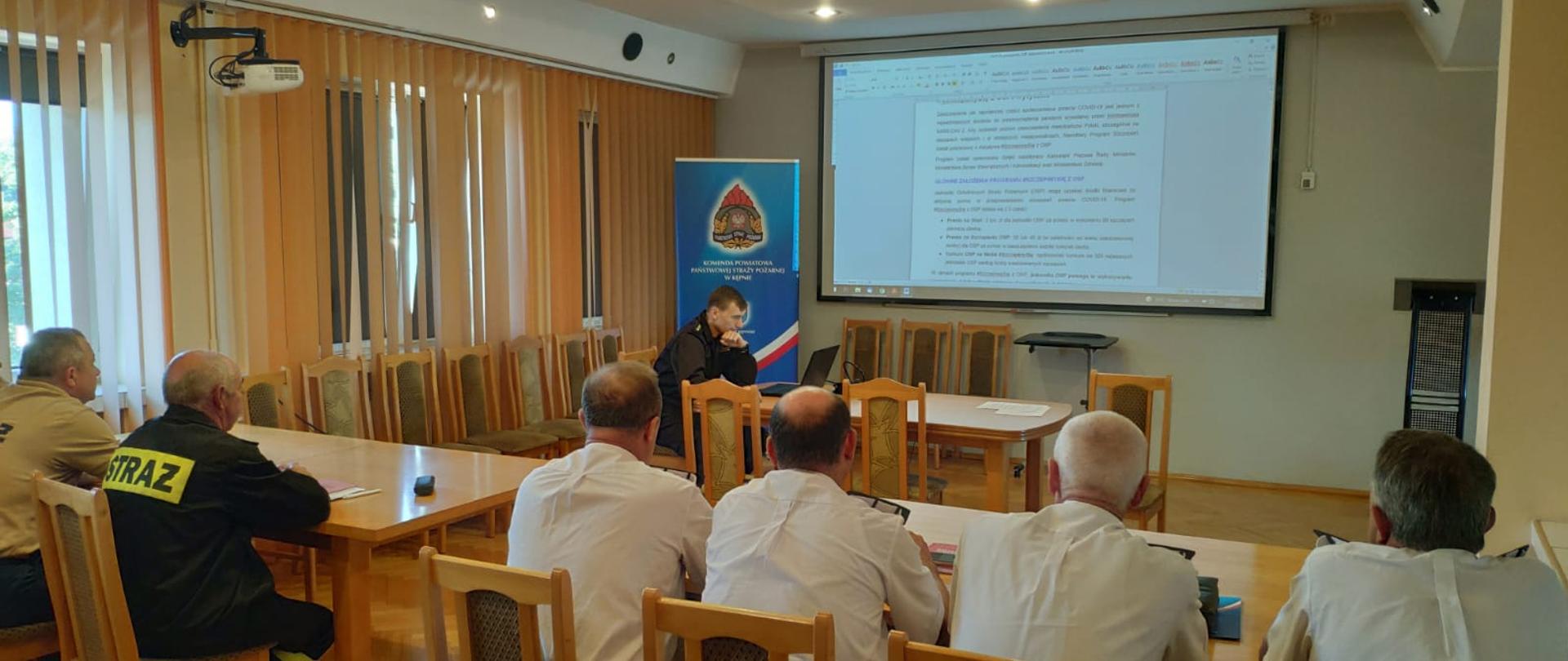 widoczni uczestnicy spotkania, druhowie OSP, oraz funkcjonariusz psp przedstawiający prezentację.