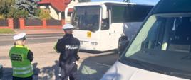 Biały autobus zaparkowany na zielonych metalowych najazdach oraz znajdujący się wokół metalowej zielonej konstrukcji z biało-zielonym logo inspekcji transportu drogowego. Trwa kontrola stanu technicznego z wykorzystaniem mobilnej stacji diagnostycznej, której przyglądają się funkcjonariuszka inspekcji transportu drogowego oraz funkcjonariusz policji. Widoczny jest również fragment radiowozu ITD. W tle widać ulicę oraz zabudowania domów jednorodzinnych. Pogodna aura z bezchmurnym niebem.