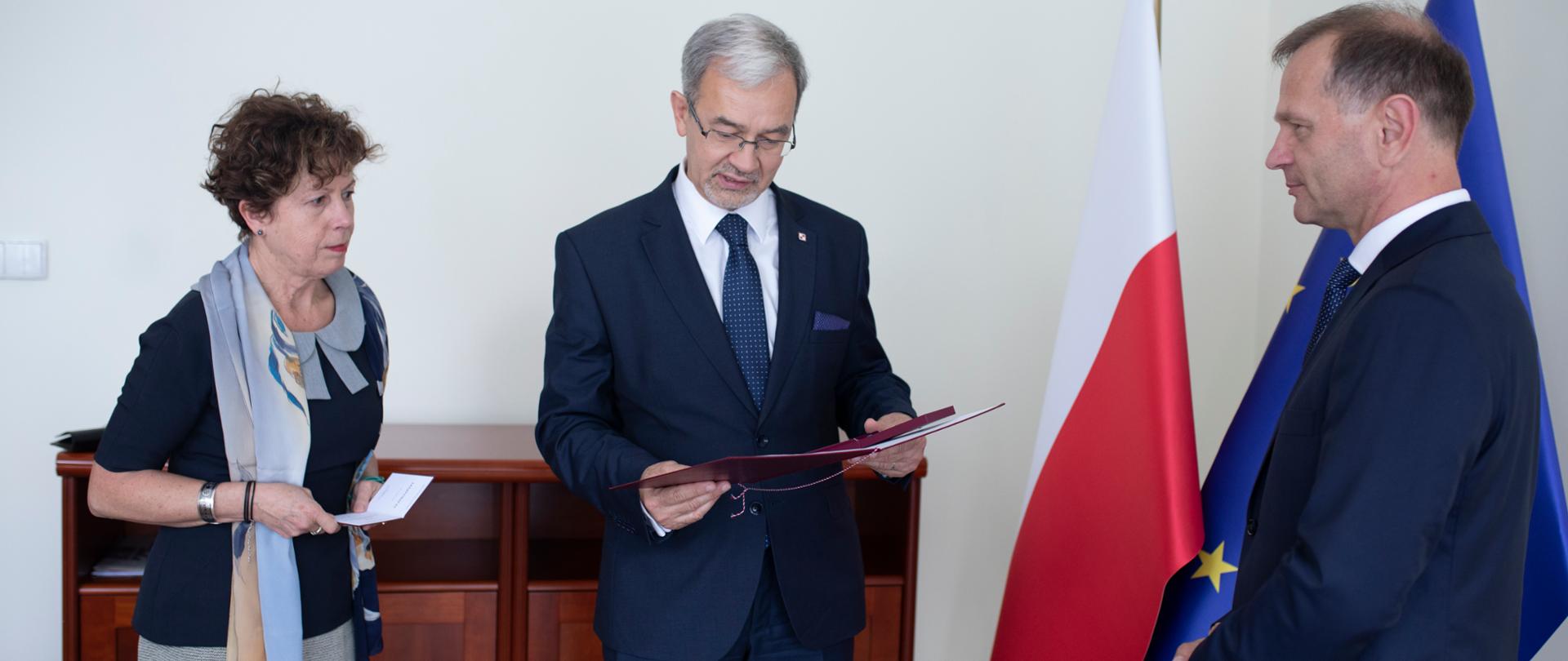 minister Jerzy Kwieciński odczytuje postanowienia o nadaniu Medalu 100-lecia Odzyskania Niepodległości, przed nim stoi uhonorowany, na zdjęciu widać też flagi polską i unijną i osobę, która podaje legitymację do odznaczenia