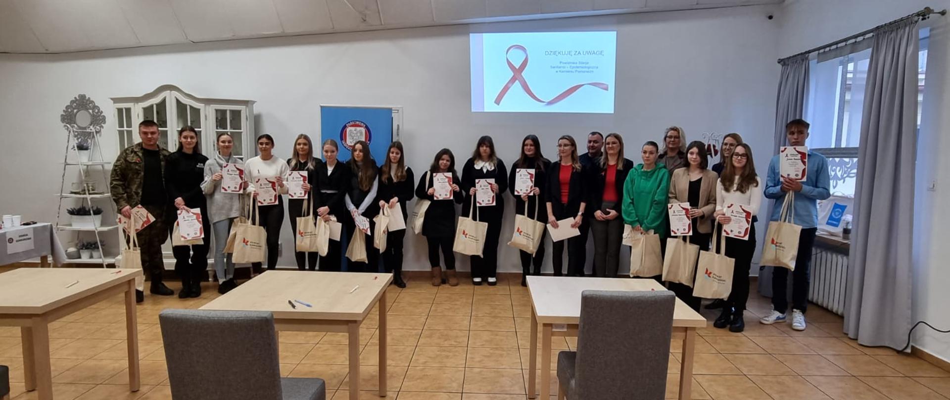 W centralnej części zdjęcia w pomieszczeniu ustawiona jest grupa ludzi - uczestnicy i organizatorzy konkursu wiedzo o HIV/AIDS. Nad nimi widoczna jest wyświetlana z projektora na ścianie czerwona kokarda - symbol Dni Walki z AIDS. Przed grupą widać kilka stanowisk do pisania testu - stoli i krzesło