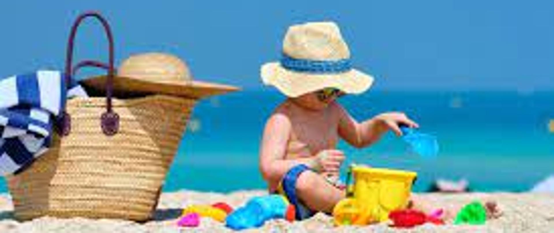 na pierwszym planie dziecko w wieku około 2 lat, ubrane w krótkie spodenki i słomiany kapelusz z niebieską obwódką. Dziecko bawi się zabawkami takimi jak łopatka do piasku w kolorze niebieskim, wiaderko w kolorze żółtym oraz drobne zabawki plastikowe. Obok dziecka na piasku stoi duża torebka w słomianym kolorze i brązowymi rączkami. na torbie leży kapelusz słomiany oraz zwinięty ręcznik w biało-niebieskie pasy. w tle błękitne morze oraz niebieskie niebo
