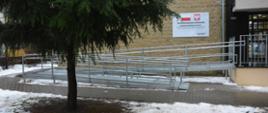 Widok na metalowy podjazd dla osób z niepełnosprawnościami przy budynku szkoły. Podjazd z poręczami prowadzi do wejścia, obok znajduje się tablica informacyjna o dofinansowaniu z Funduszu Promocji Kultury. Na pierwszym planie drzewo oraz fragment chodnika oraz śnieg