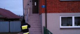 Strażacy roznoszą ulotki do gospodarstw domowych na terenie powiatu ostrowieckiego. Na zdjęciu widać strażaka przed domem mieszkalnym, który zostawia broszurę informacyjną.
