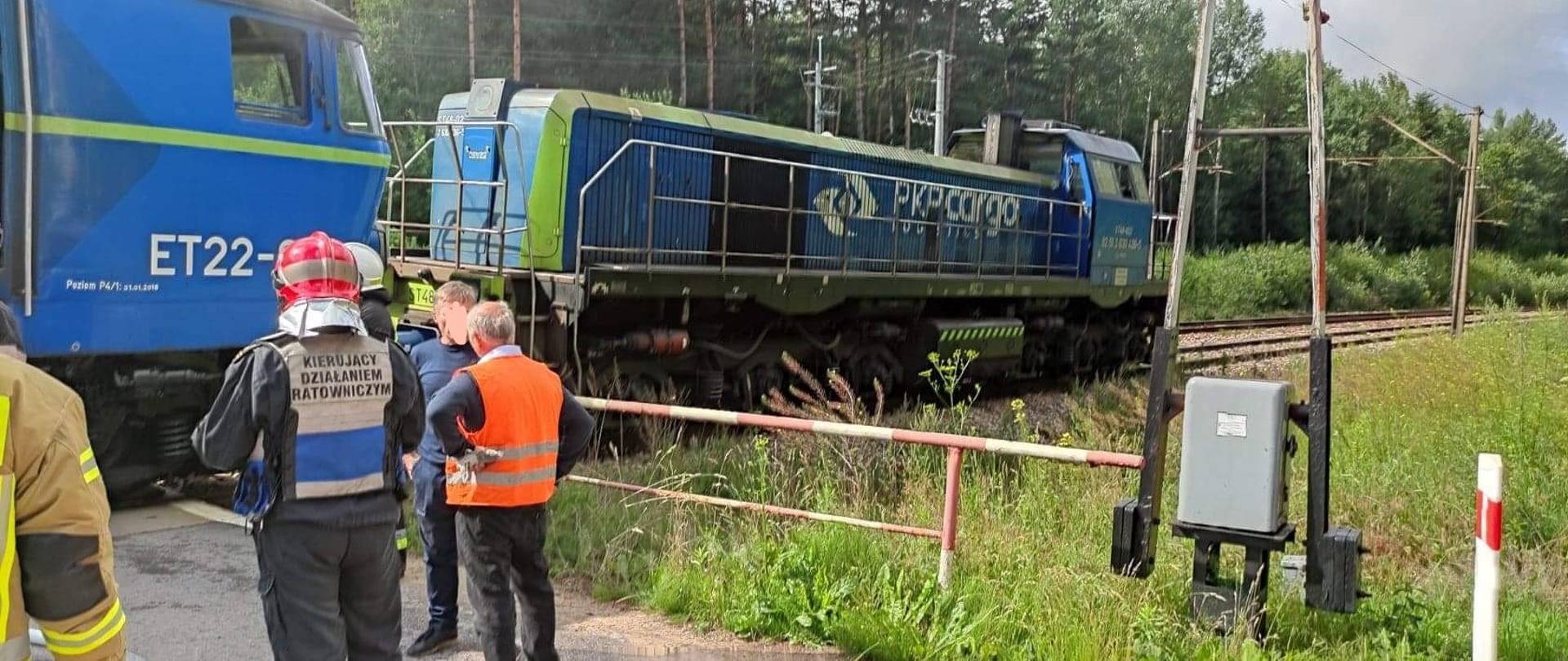 Na zdjęciu widać strażaków i służby PKP przy przejeździe kolejowym stojących przy lokomotywach towarowych. W tle widać drzewa. 