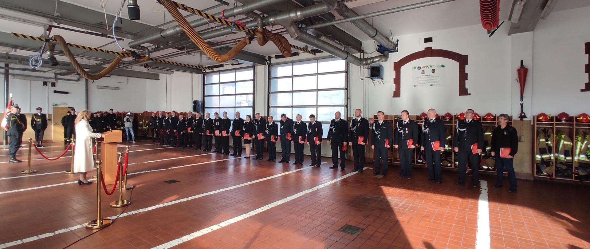 Przemówienie Poseł na Sejm RP Pani Katarzyny Czochary podczas uroczystego wręczenie promes dla Młodzieżowych Drużyn Pożarniczych OSP powiatu prudnickiego. Na zdjęciu oprócz przemawiającej widoczni przedstawiciele jednostek OSP powiatu prudnickiego, którzy otrzymali promesy. Strażacy OSP w ubraniach wyjściowych.