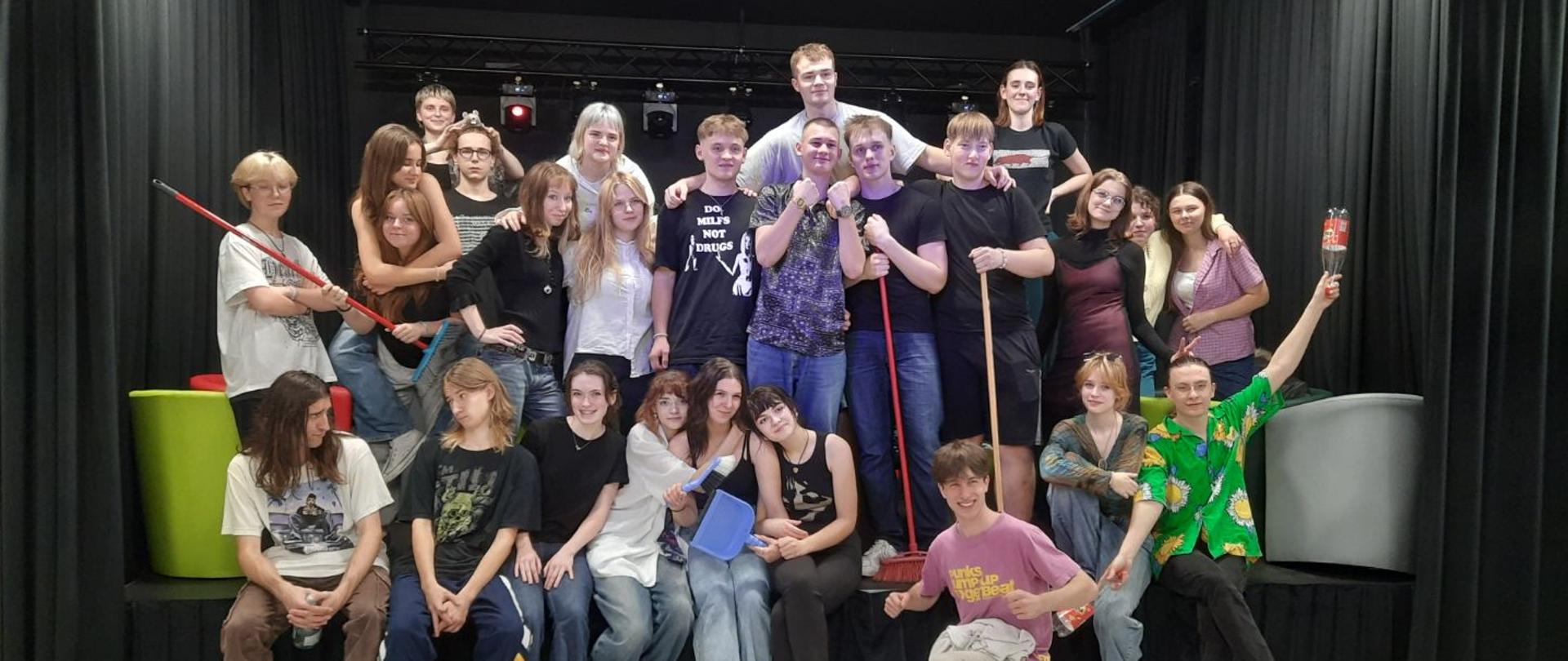 Kilkunastoosobowa grupa młodzieży pozuje do zdjęcia, część z nich siedzi, część stoi na scenie w kotłowni artystycznej. Ubrani na kolorowo, kilka z nich trzyma przyrządy do sprzątania, chłopiec siedzący po prawej stronie trzyma butelki po oranżadzie